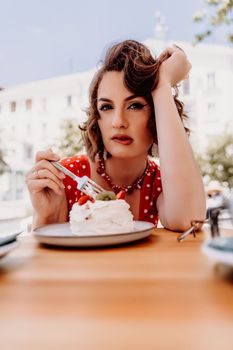 Charming woman in a restaurant, cafe on the street. She sits at the table and eats a cake with a fork. Dressed in a red sundress with white polka dots