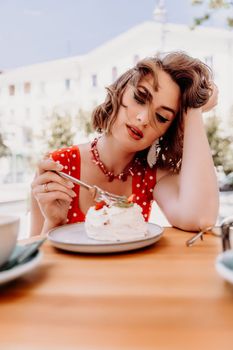 Charming woman in a restaurant, cafe on the street. She sits at the table and eats a cake with a fork. Dressed in a red sundress with white polka dots