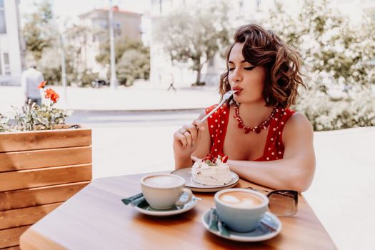 Charming woman in a restaurant, cafe on the street. She sits at the table and eats a cake with a fork. Dressed in a red sundress with white polka dots