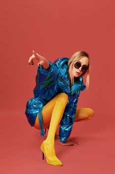 Photo of young cheeky confident blonde girl in blue jacket squats in studio with one leg bent forward before camera on red background. She wears yellow stockings and sunglasses