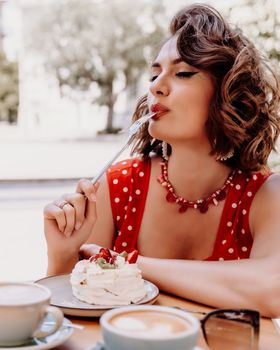 Charming woman in a restaurant, cafe on the street. She sits at the table and eats a cake with a fork. Dressed in a red sundress with white polka dots