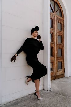 Stylish woman in the city. Fashion photo of a beautiful model in an elegant black dress posing against the backdrop of a building on a city street.