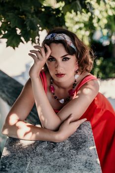 A pretty woman in a red silk dress and a bandage on her head smiles against the background of the leaves of a tree. She is leaning on the coop and looking into the camera. Vertical photo