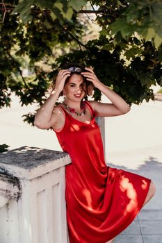 A pretty woman in a red silk dress and a bandage on her head smiles against the background of the leaves of a tree. She is leaning on the coop and looking into the camera. Vertical photo