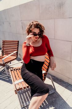 Portrait of a woman on the street. An attractive woman in glasses, a red blouse and a black skirt is sitting on a bench outside