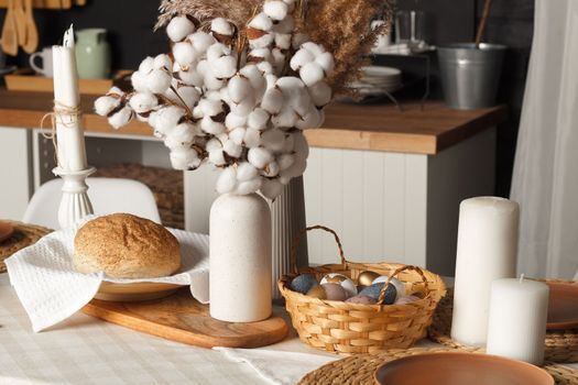 Painted eggs in a basket and bread lie on a table decorated with Easter decor. In the background is a white Scandinavian style kitchen. The concept of home comfort and decor for the Easter holiday