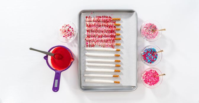 Flat lay. Drizzling melted chocolate over chocolate-dipped pretzels rods and decorating with sprinkles to make chocolate-covered pretzel rods for Valentine's Day.