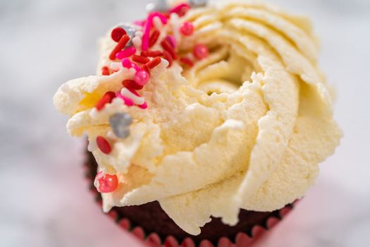 Piping white chocolate ganache frosting on top of red velvet cupcakes and topping with sprinkles.