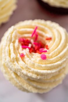Freshly baked velvet cupcakes with white chocolate ganache frosting decorated with sprinkles.