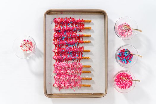 Flat lay. Drizzling melted chocolate over chocolate-dipped pretzels rods and decorating with sprinkles to make chocolate-covered pretzel rods for Valentine's Day.