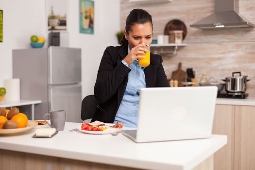 Woman enjoying an orange juice in the morning during a video conference. Concentrated business woman in the morning multitasking in the kitchen before going to the office, stressful way of life, career and goals to meet.
