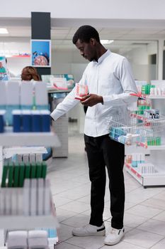 African american man checking heart nutritional supplements in drugstore, reading instruction on vitamins package. Buyer choosing medicaments, buying medications, wide shot