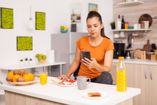 Woman checking social media in the morning at breakfast time. Housewife using modern tehcnology and drinking healthy, natural, homemade orange juice. Refreshing morning.