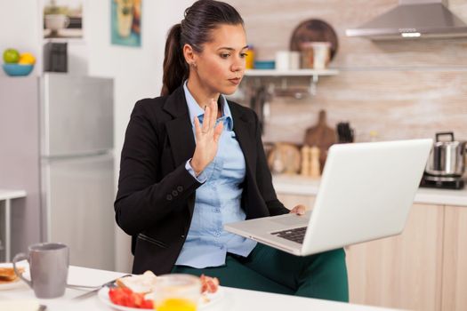 Business woman waving on video call during breakfast. Young freelancer in the kitchen having a healthy meal while talking on a video call with her colleagues from the office, using modern technology and working around the clock