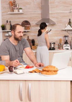 Entrepreneur working from home while eating breakfast in kitchen, wearing pajamas enjoying roasted bread with butter. Freelancer working online via internet using modern digital technology