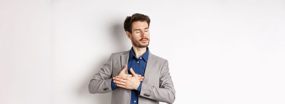 Romantic bearded man in stylish suit holding hands on heart, close eyes and remember something with tender feelings, standing nostalgic on white background.