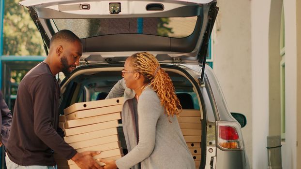 African american courier giving piles of pizza boxes to diverse people standing near building entrance. Food delivery employee delivering stacks of meal packages in car trunk. Tripod shot.