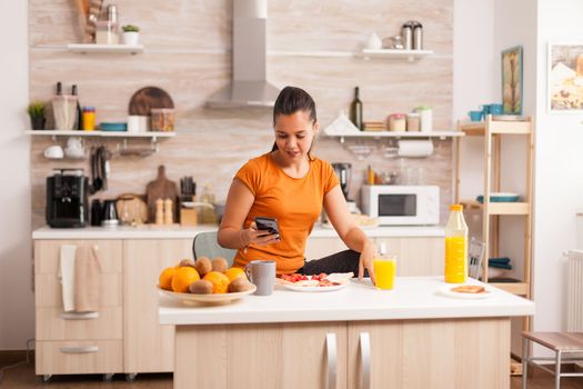 Freelancer reading news on phone in kitchen during breakfast. Housewife using modern tehcnology and drinking healthy, natural, homemade orange juice. Refreshing morning.