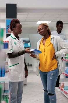 Customer buying back pain medication in drugstore, pharmacy consultant and buyer talking. African american woman client with backache standing, shopping, chatting with pharmacist, all black team