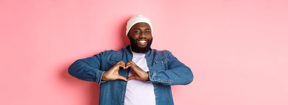 Happy african-american man showing heart sign, I love you gesture, smiling at camera, pink background.