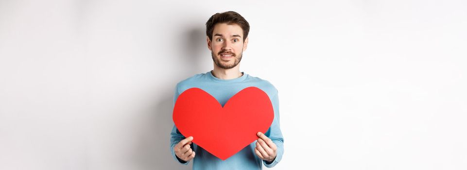 Relationship and love concept. Handsome caucasian man in sweater holding big red valentines day heart cutout and smiling, confessing on date, standing over white background.