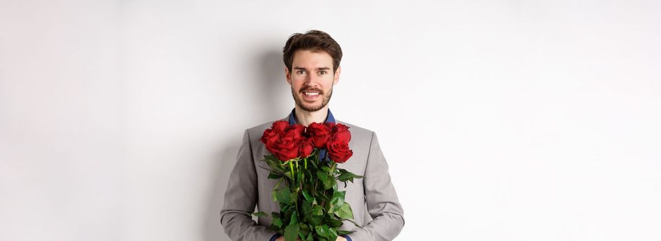 Handsome young man in suit standing with gift on valentines day, holding red roses bouquet and smiling at lover, standing over white background.