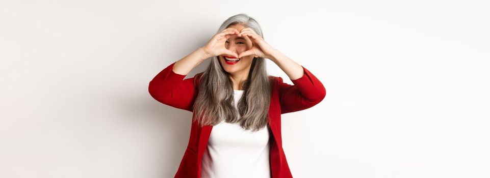Lovely asian middle-aged woman with grey hair, wearing red blazer, showing heart sign and peeking throught it, I love you gesture, standing over white background.