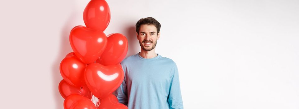 Smiling young man standing with heart balloons and looking happy, celebrating valentines day, bring romantic present to lover, standing over white background.