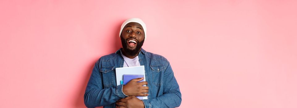 Education. Happy african-american male student in beanie holding notebooks, studying courses, smiling at camera, standing over pink background.