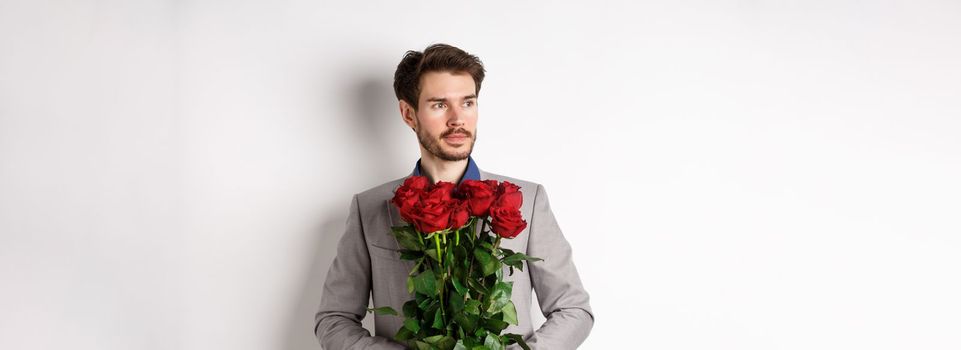 Handsome boyfriend in suit going on romantic date, holding bouquet of red roses and looking left thoughtful, standing over white background.