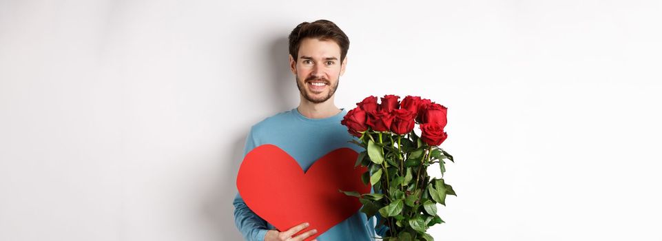 Romantic people. Handsome smiling man holding bouquet of roses and big red heart, going on Valentines day date with girlfriend, standing over white background.