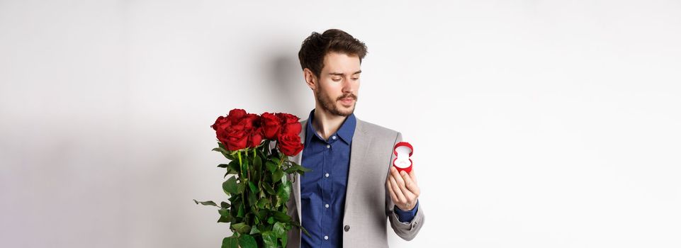 Handsome bearded man in suit looking at engagement ring, making surprise on lovers day, standing with red roses over white background.