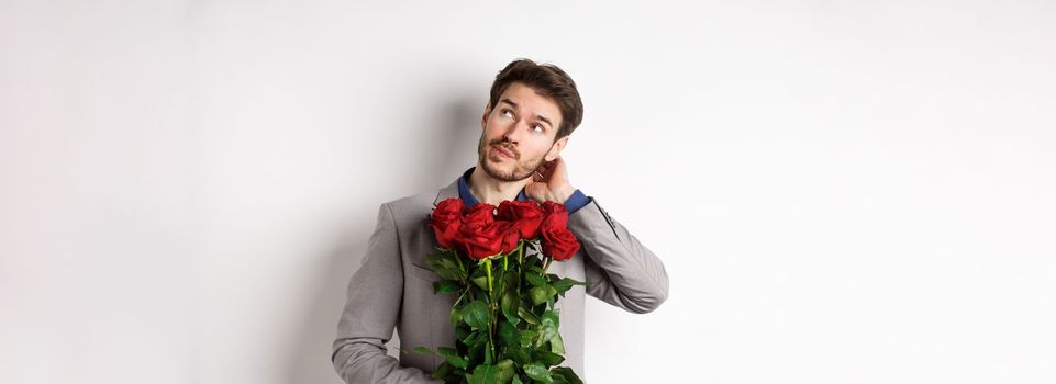 Thoughtful young man in suit going on valentines day date, holding bouquet of roses, thinking and scratching head, standing against white background.