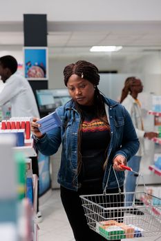 African american customer buying medication in pharmacy, reading instruction, holding tablets package, standing in drugstore aisle. Client choosing vitamins, carrying basket, medium shot