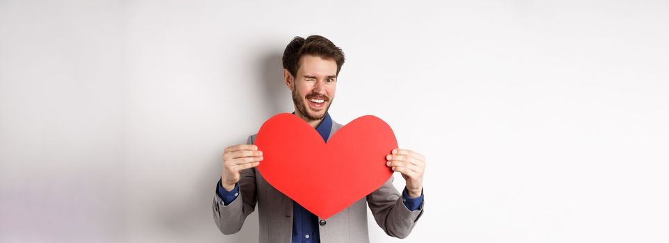 Charismatic young man winking and smiling, showing big red heart cutout for Valentines day date, say love you to lover, standing over white background.
