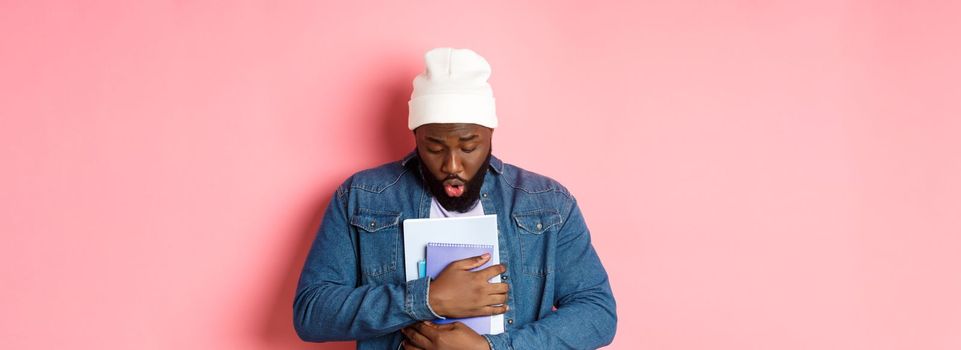 Education. Image of african-american bearded male student holding notebooks and looking down, drop something on floor, standing over pink background.