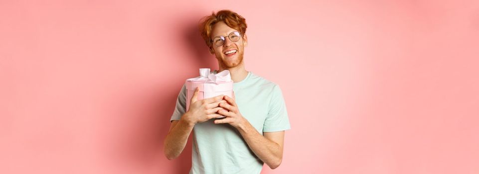 Valentines day and romance concept. Happy redhead boyfriend receive romantic gift, hugging box with present and saying thank you, smiling grateful, pink background.