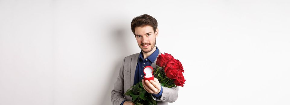 Romantic man with boquet of red roses asking to marry him, holding engagement ring and looking confident at camera, standing in suit over white background.