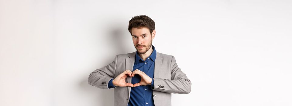 Romantic man in suit showing heart sign and smiling, love his girlfriend, standing on white background.