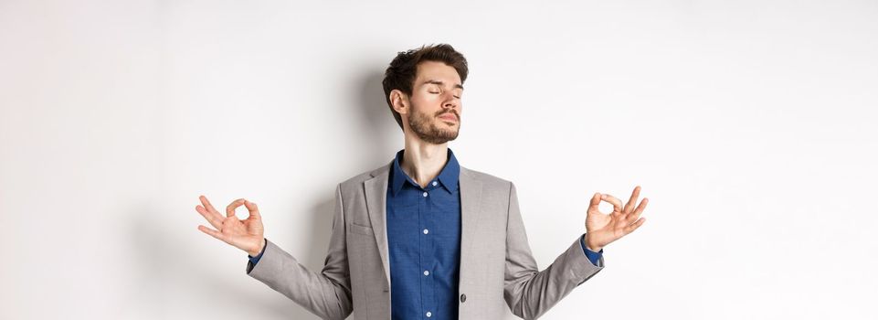 Calm and focused businessman meditating with eyes closed and hands spread sideways, finding peace in meditation, practice yoga breathing, standing on white background.