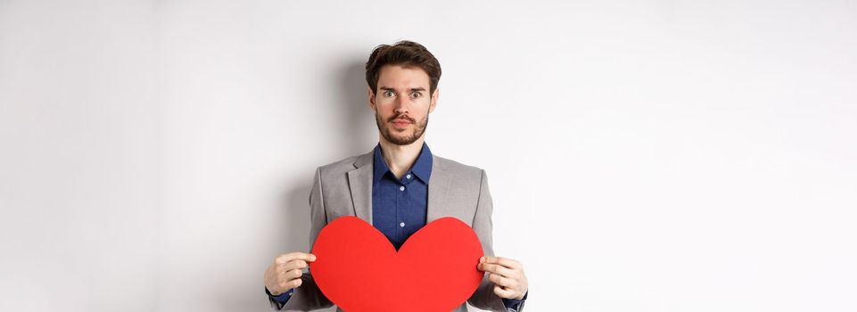 Excited caucasian man in suit looking at camera, holding big red heart cutout on valentines day, standing over white background. Copy space