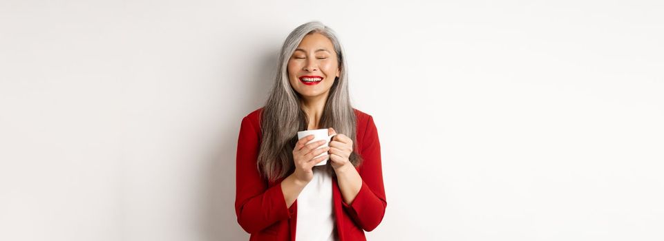 Business people concept. Happy office lady in red blazer enjoying drinking coffee, holding mug and smiling delighted, standing over white background.