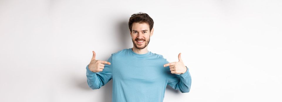 Handsome bearded man pointing fingers at center, self-promoting and looking confident, standing over white background, smiling at camera.