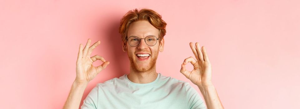 Headshot portrait of attractive young man with messy red hair and beard, wearing glasses, smiling with white teeth and showing Ok signs in approval, praise something good.