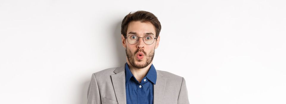 Close up portrait of excited office worker in glasses and suit saying wow, staring amazed at camera, standing against white background.