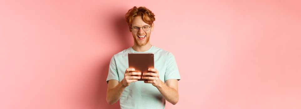 Happy guy with red hair and beard using digital tablet, reading screen and smiling, standing over pink background.