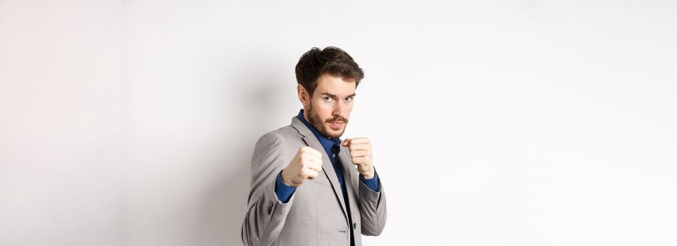 Serious man in suit raising hands in boxer pose, going to fight, looking confident, beckon to attack, standing on white background in defensive pose.