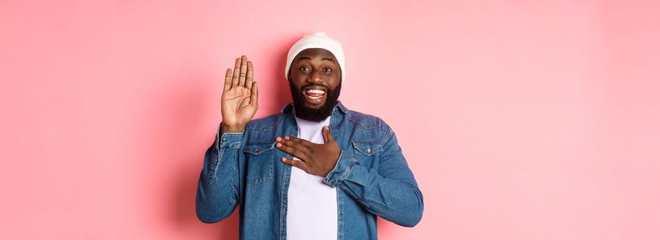 Image of smiling african-american man being honest, telling truth, hands on heart and in air, making promise or swearing, standing ove rpink background.