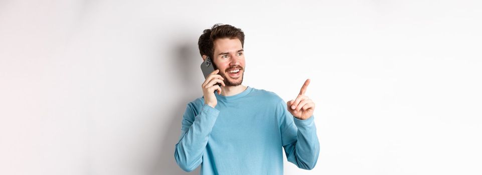 Technology concept. Young man talking on mobile phone and pointing left, having call on smartphone, standing over white background.