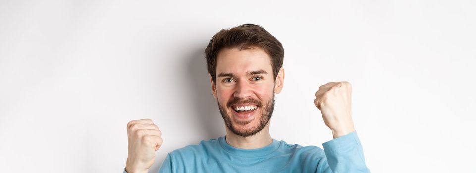 Close-up of happy young man celebrating, winning prize and say yes, smiling satisfied with fist pumps, standing over white background.